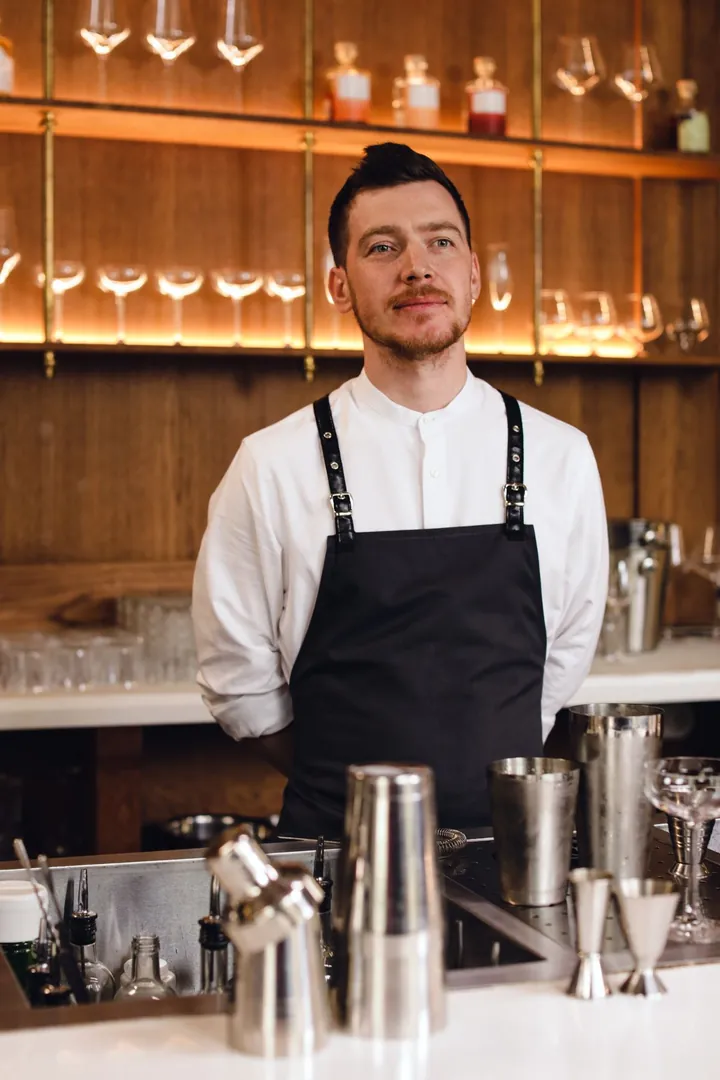 Barman en chemise blanche et tablier à bavette noir à bretelles de cuir derrière un comptoir garni de shakers